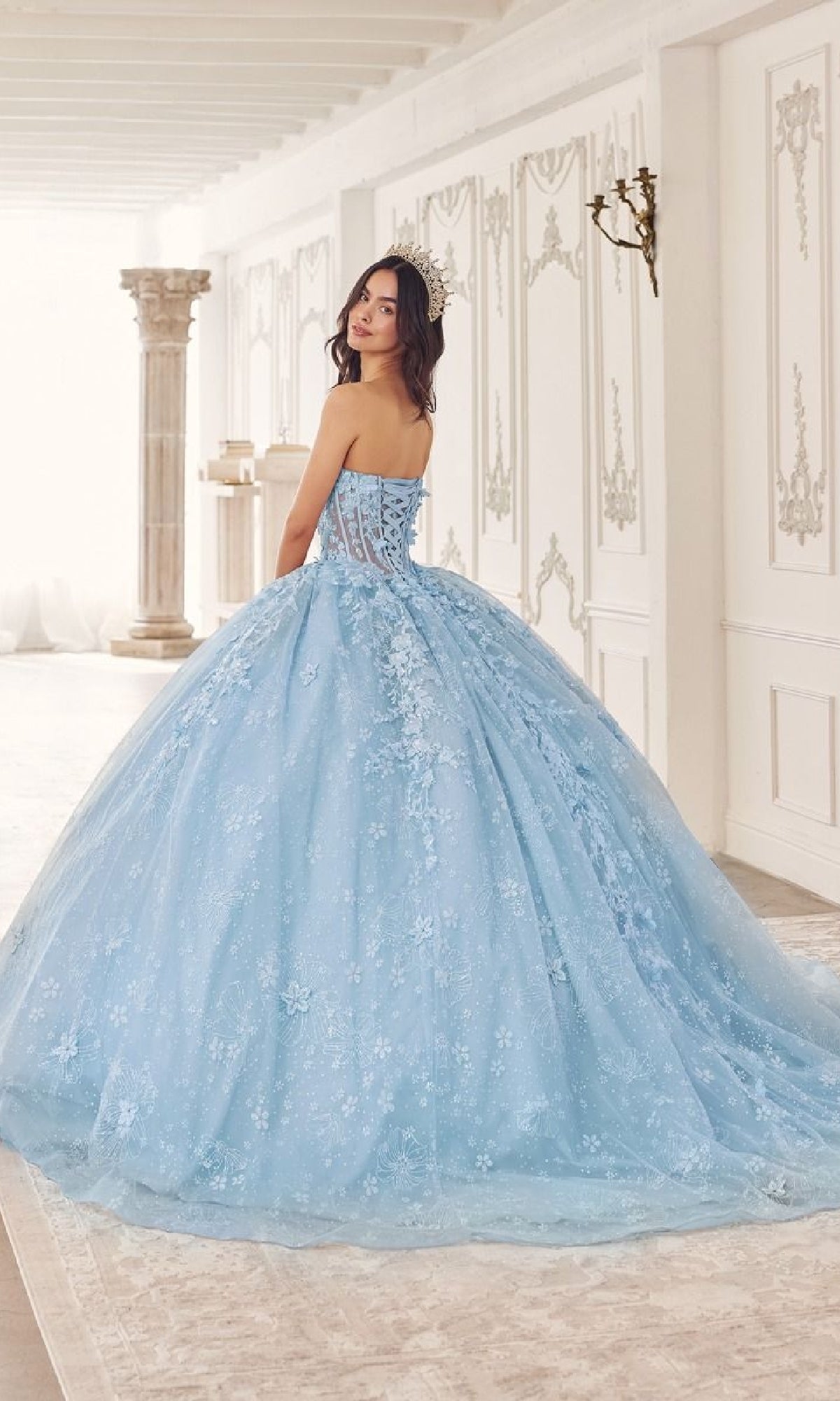 A woman in the Quinceanera Dress 15719 by Ladivine, a strapless ball gown with floral patterns, stands in an elegant white classical room, looking over her shoulder.